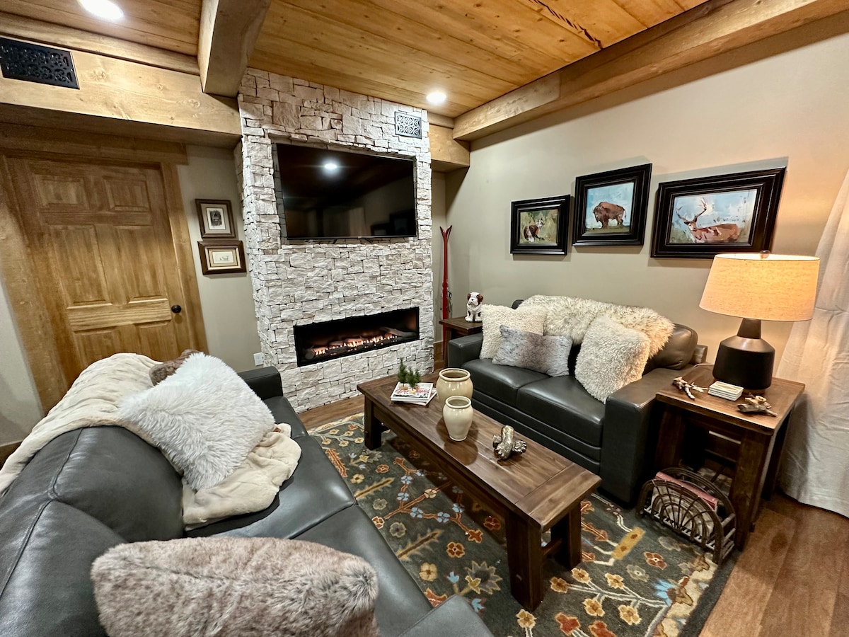 A cozy living space features a rustic stone fireplace and a mounted television above it. Dark leather sofas are adorned with soft throws and cushions. A wooden coffee table is decorated with neutral-toned decor items, and framed wildlife artwork hangs on the wall.