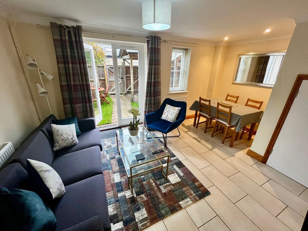 A spacious living area features a dark sofa paired with a geometric coffee table. Natural light enters through sliding glass doors, leading to the courtyard garden. An adjacent dining area is furnished with a wooden table and six chairs, complemented by patterned decor and soft lighting.