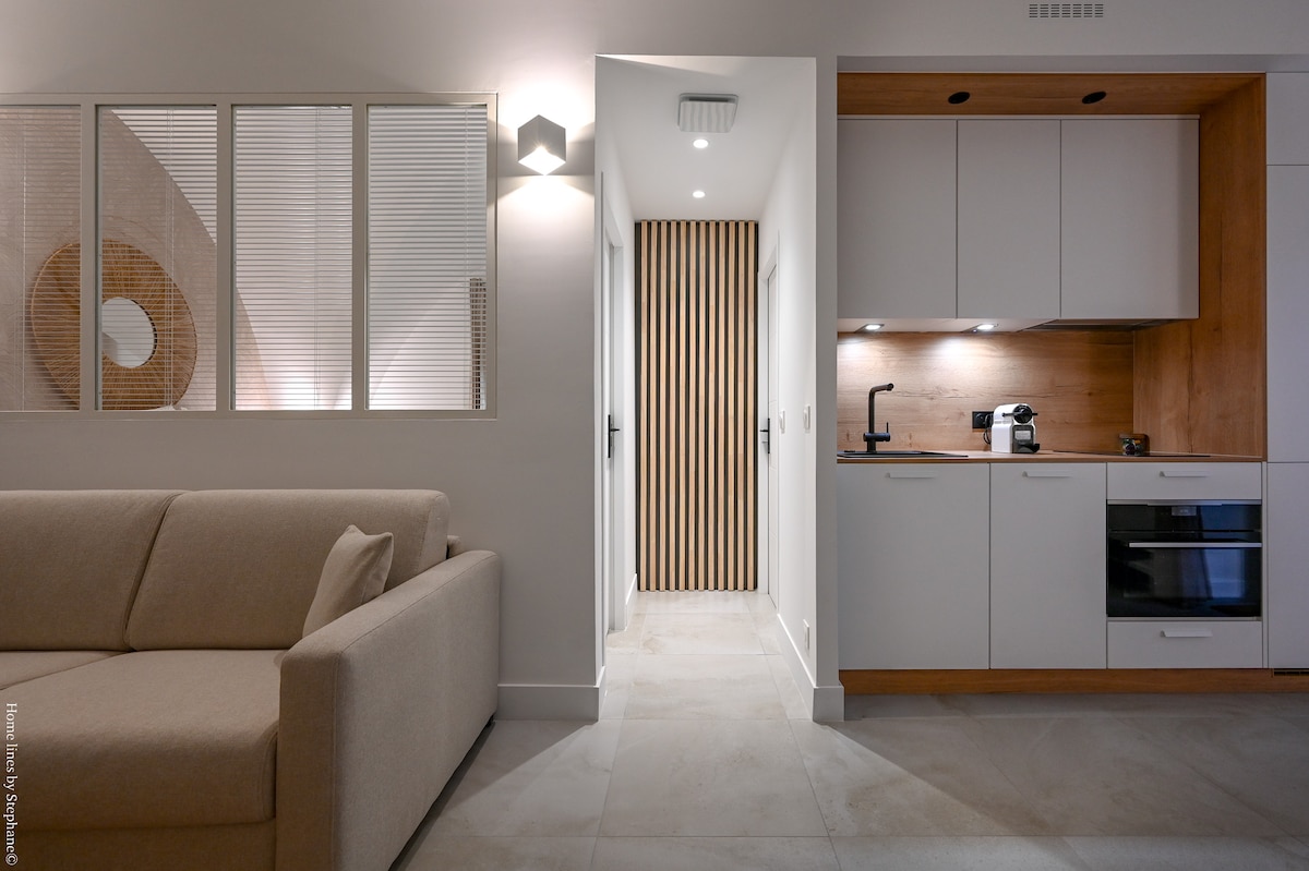 A modern living area features a light-colored couch positioned against a wall. The space transitions into a kitchen area with wood accents and sleek appliances. Soft lighting is provided by a wall fixture, and a hallway leading to additional rooms is visible in the background.
