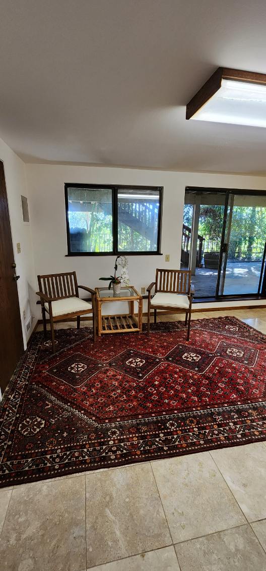 A seating area is showcased with two wooden chairs arranged around a small table. A decorative rug with rich colors lies beneath, while large windows allow natural light to illuminate the space, revealing views of the outdoor greenery.