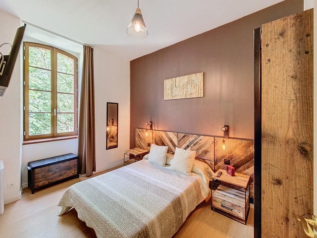 A cozy bedroom features a spacious bed with neutral bedding, complemented by two decorative cushions. A wooden headboard adds character, and a framed artwork is displayed above. Natural light filters in through a large window, while a wall-mounted television and a chest are positioned thoughtfully within the space.