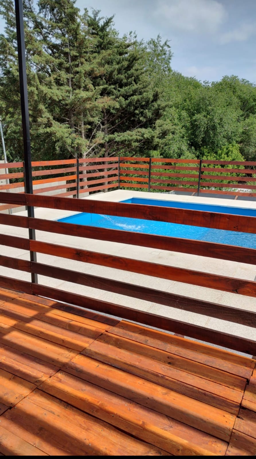 A wooden deck surrounds a rectangular pool, enhanced by the natural greenery in the background. Vertical wooden slats create a stylish barrier, allowing glimpses of the water while providing privacy. Soft sunlight illuminates the scene, creating a serene outdoor space.
