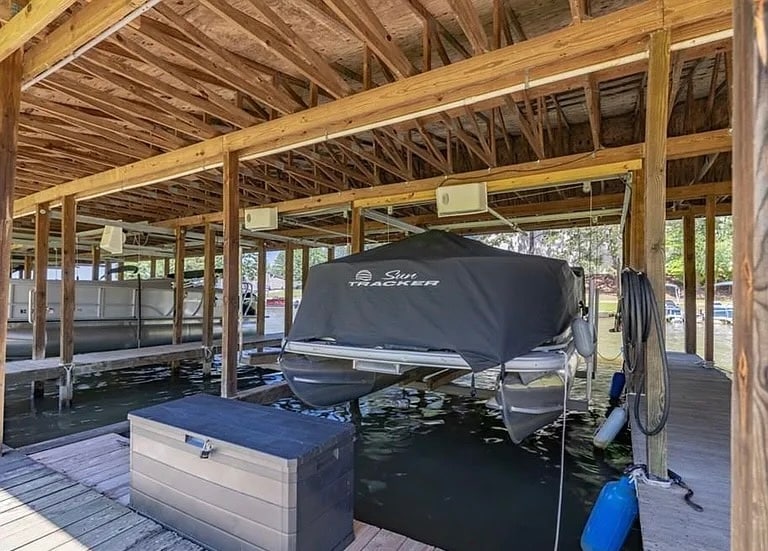 A covered boat slip features a stored pontoon boat secured under a protective cover. The wooden structure is supported by strong beams, with a visible dock area extending outward into the lake. Water reflections create a serene setting, inviting guests to enjoy water activities.