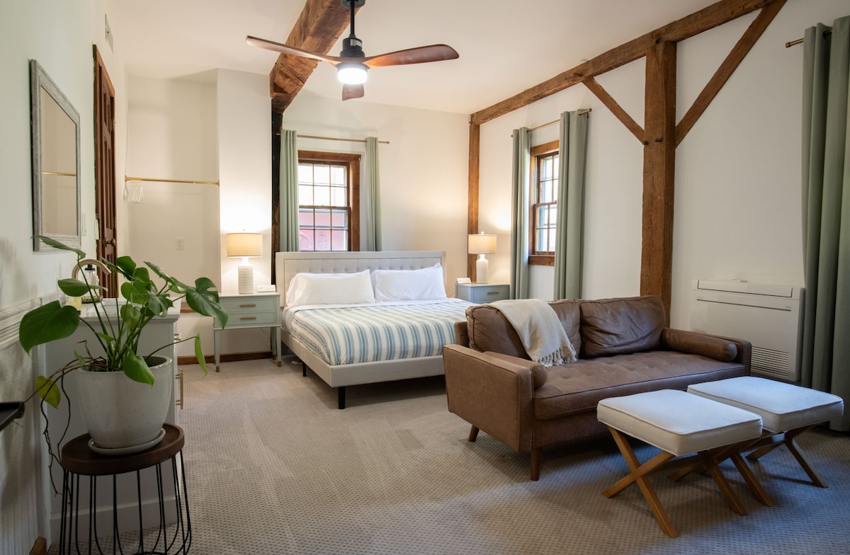 A spacious room features a king bed with a light-colored striped blanket, complemented by a comfortable sitting area that includes a brown sofa and two white stools. Large windows provide natural light, while wooden beams add character to the ceiling.
