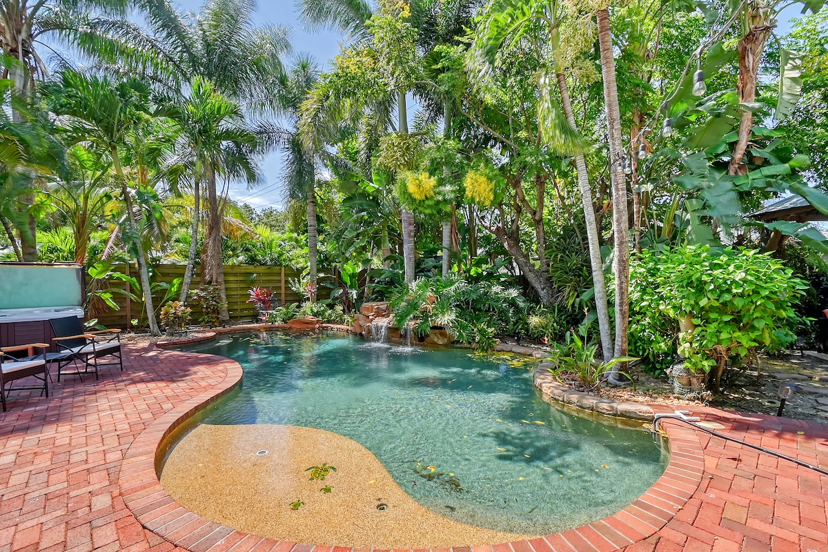 A saltwater pool is surrounded by lush tropical plants and palm trees, creating a private oasis. A gently flowing waterfall enhances the natural setting, and a paved area borders the pool, providing space for relaxation and outdoor enjoyment.