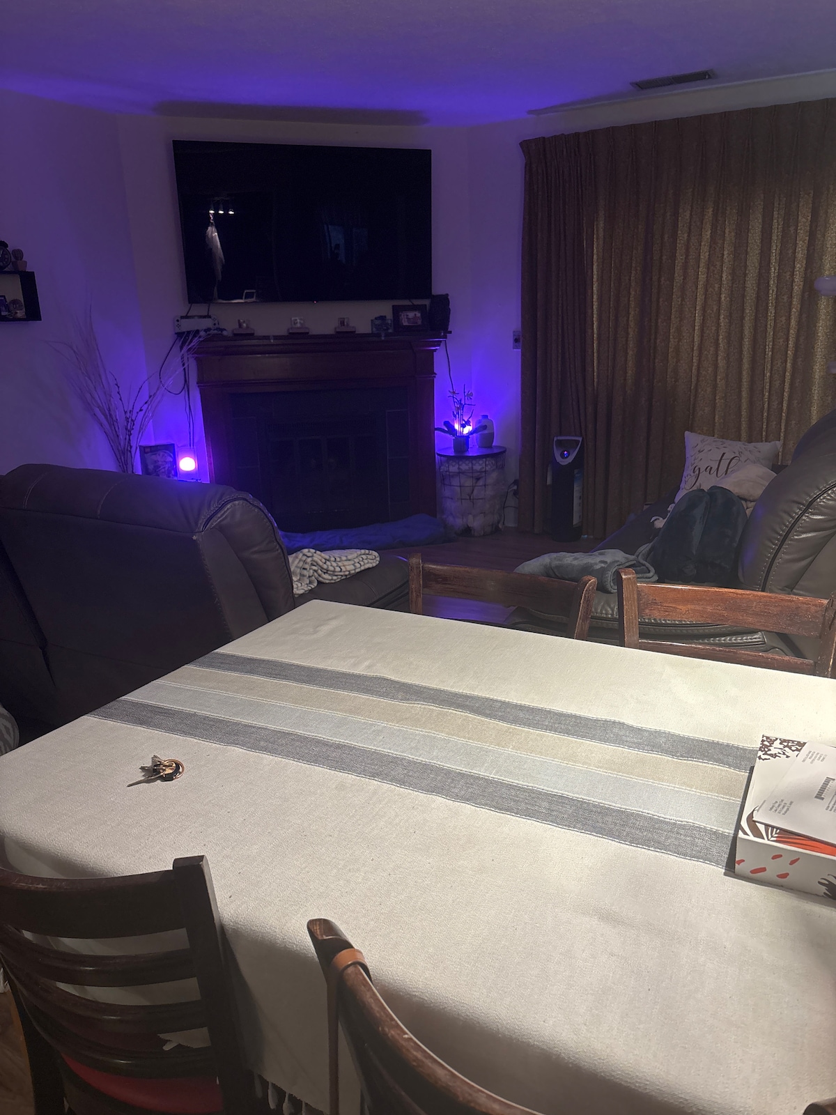 A dining area is highlighted by a light-colored table covered with a striped cloth, surrounded by wooden and upholstered chairs. A cozy seating space is visible in the background, accented by soft lighting and a fireplace, creating an inviting space for relaxation.