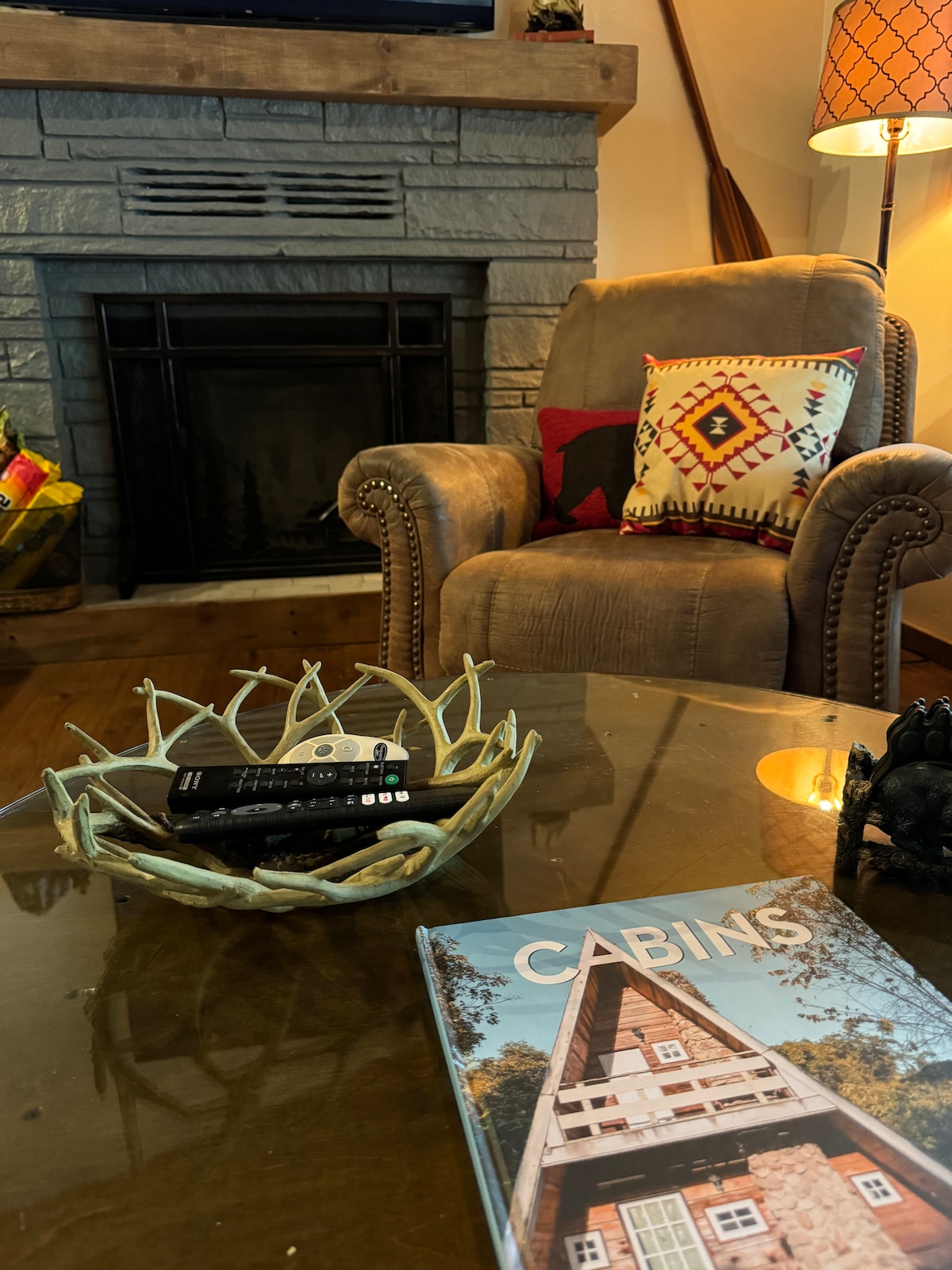 A comfortable armchair is positioned beside a fireplace, featuring a stone surround. A decorative bowl with a remote control rests on a glass table, which also holds a magazine displaying a cabin on its cover. Soft lighting is provided by a nearby lamp.