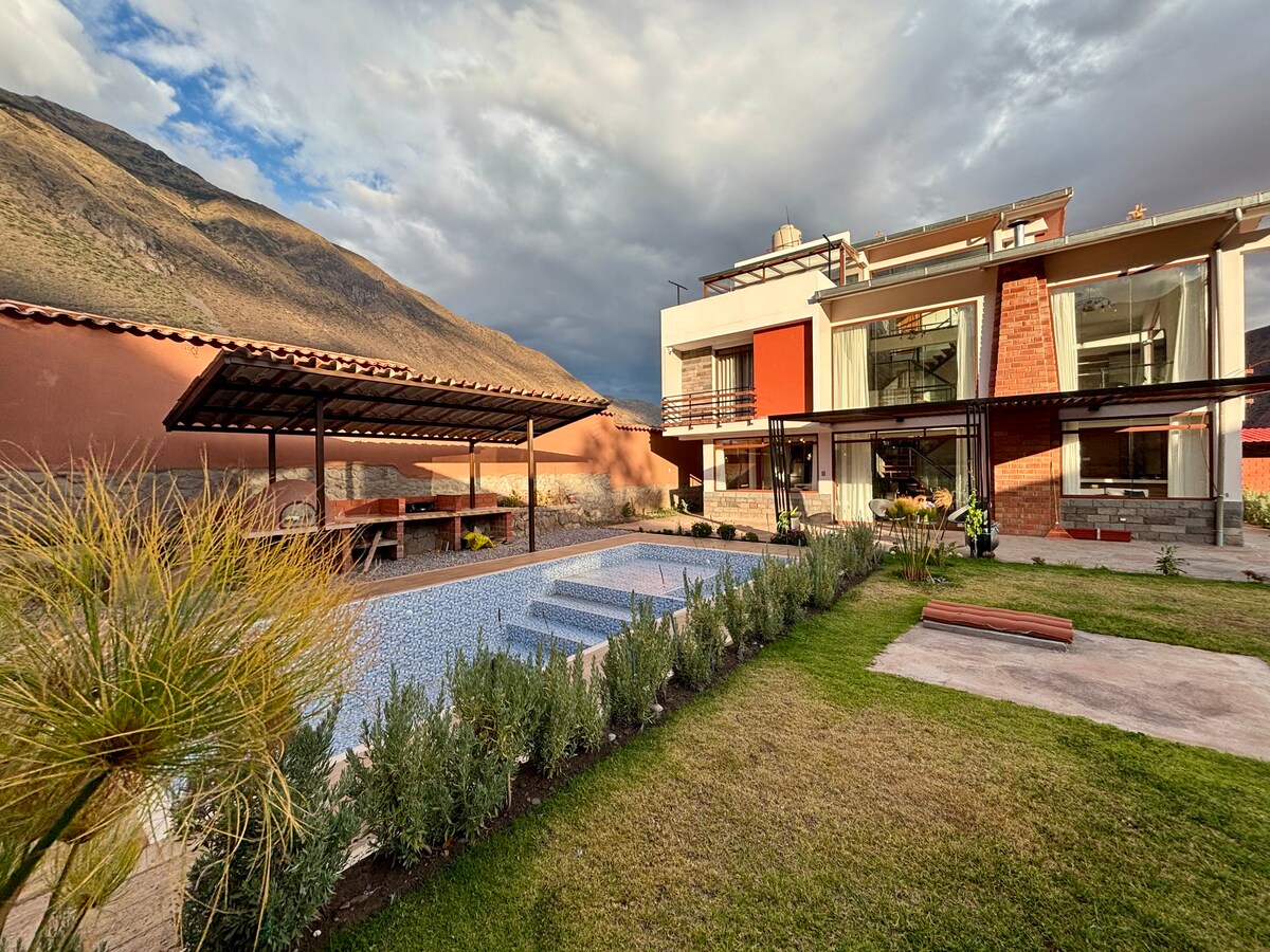 An inviting outdoor area showcases a well-maintained garden with neatly arranged plants and a swimming pool. The building features large glass windows and a covered patio with seating, framed by scenic mountain views under a partly cloudy sky.