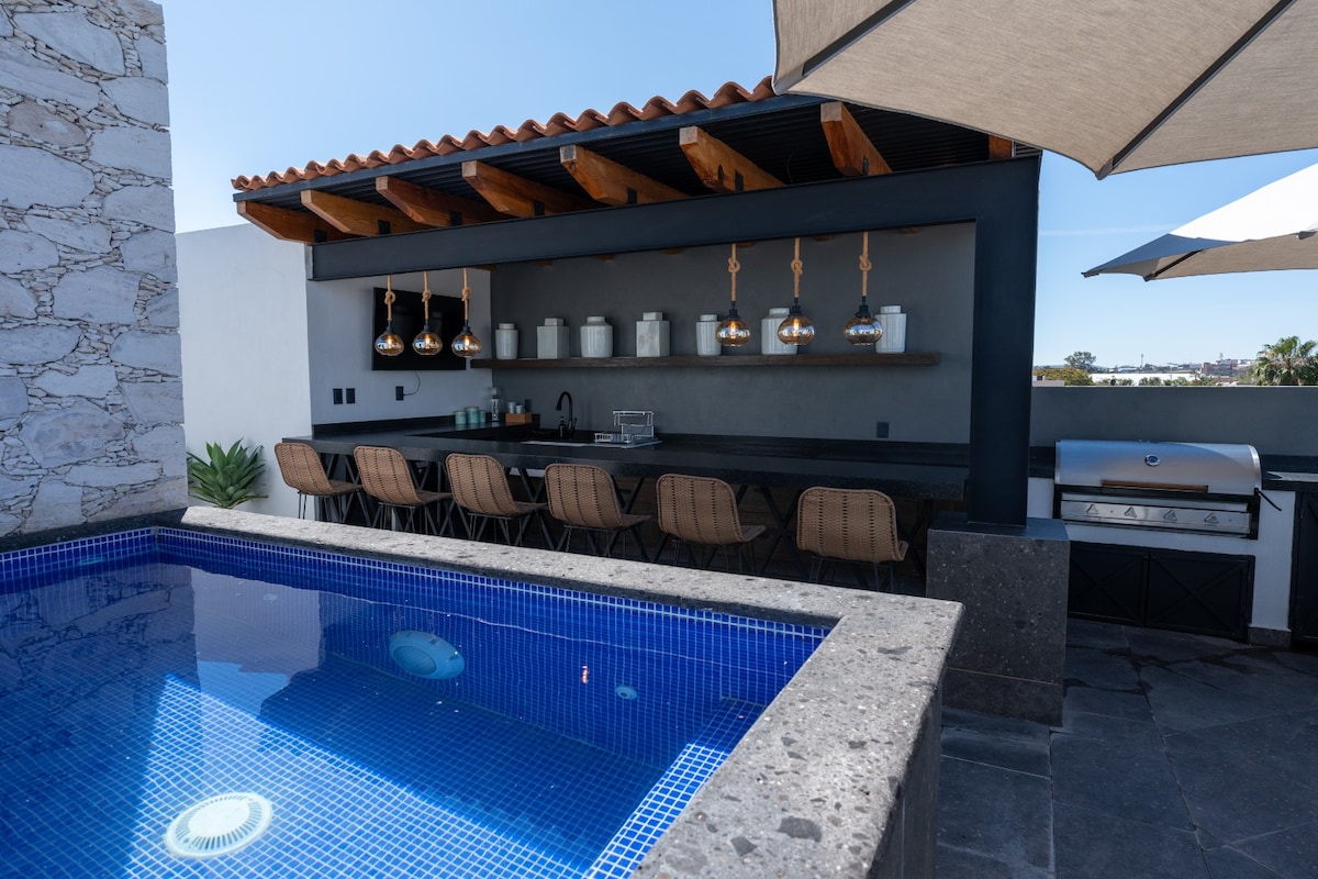 A private rooftop area features a blue tiled jacuzzi and an outdoor kitchen with metallic pendant lights and high-backed seating. A grill is positioned beside the kitchen. Sun umbrellas provide shade, and the space is surrounded by a wall that enhances privacy.
