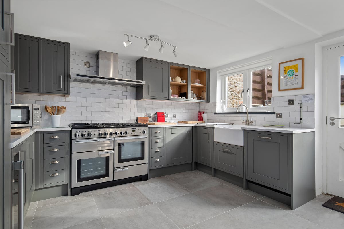The modern kitchen features an L-shaped layout with grey cabinetry and stainless steel appliances. A large farmhouse sink is set against the wall beneath a window, while a spacious countertop provides ample workspace. Natural light enhances the clean lines and functional design.