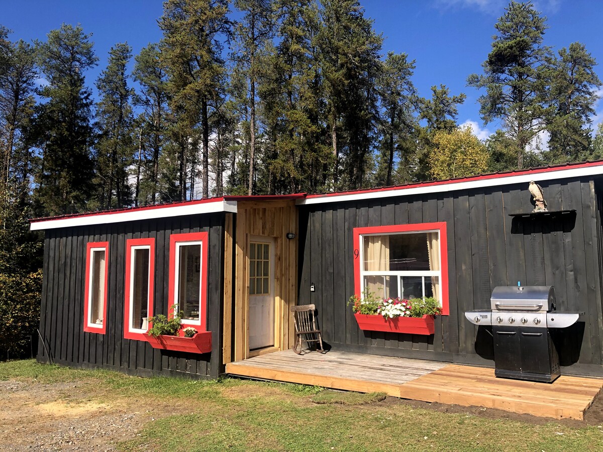 The exterior of the cottage features a dark wooden facade with red trim around the windows. A wooden deck extends from the entrance, which is flanked by two flower boxes. A grill is situated on the deck, surrounded by grassy landscaping and tall trees in the background.