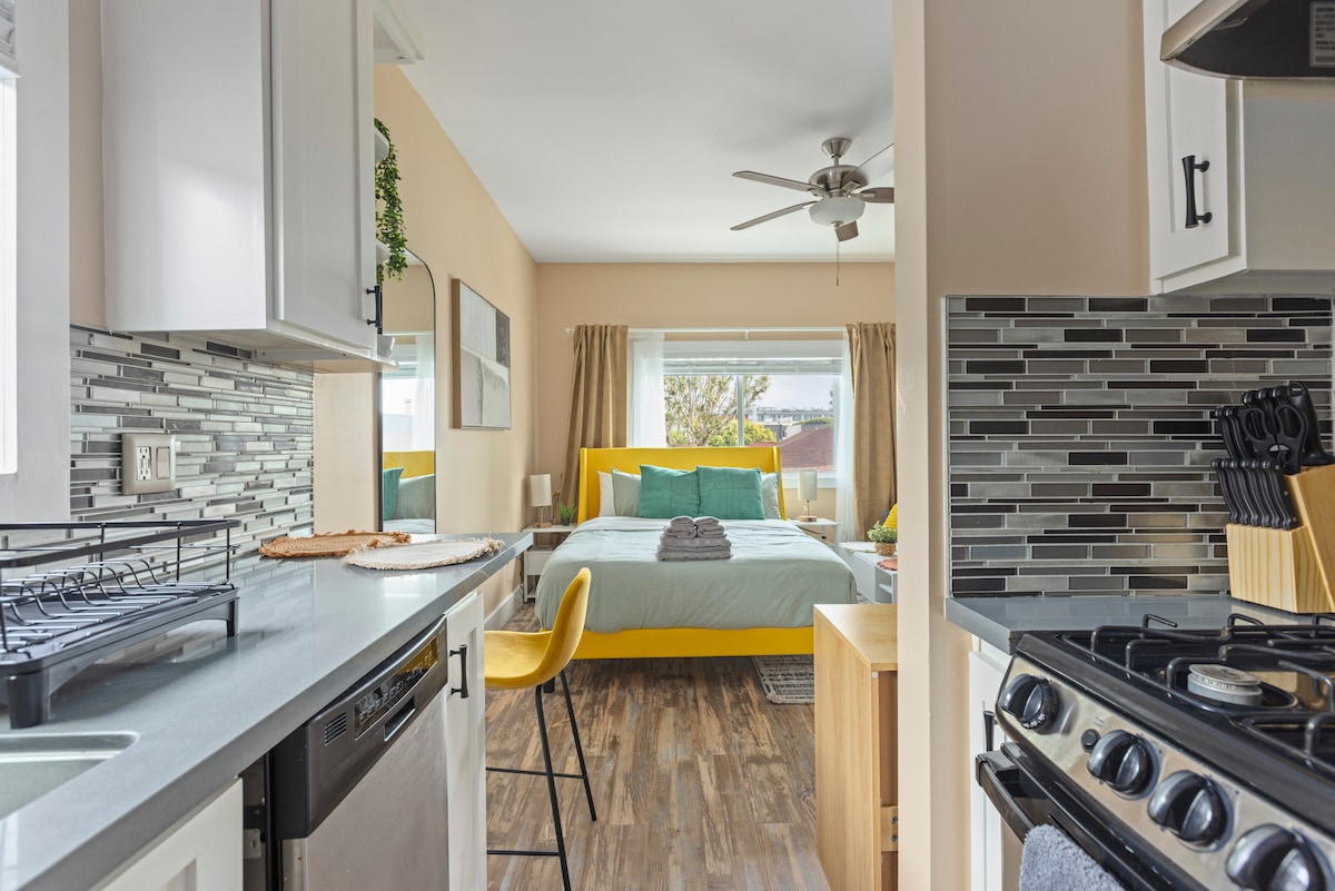 A spacious studio layout is showcased, featuring a fully equipped kitchen with modern appliances on the left. The inviting sleeping area, set against yellow accents, offers a cozy queen bed with light-blue bedding. Natural light filters through large windows, enhancing the warm atmosphere.