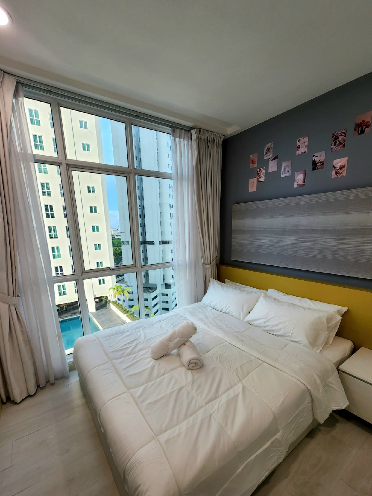 A cozy bedroom features a spacious bed dressed in white linens, accompanied by a rolled towel. Large windows provide natural light and a view of the surrounding buildings, while soft grey and yellow accents create a soothing atmosphere.