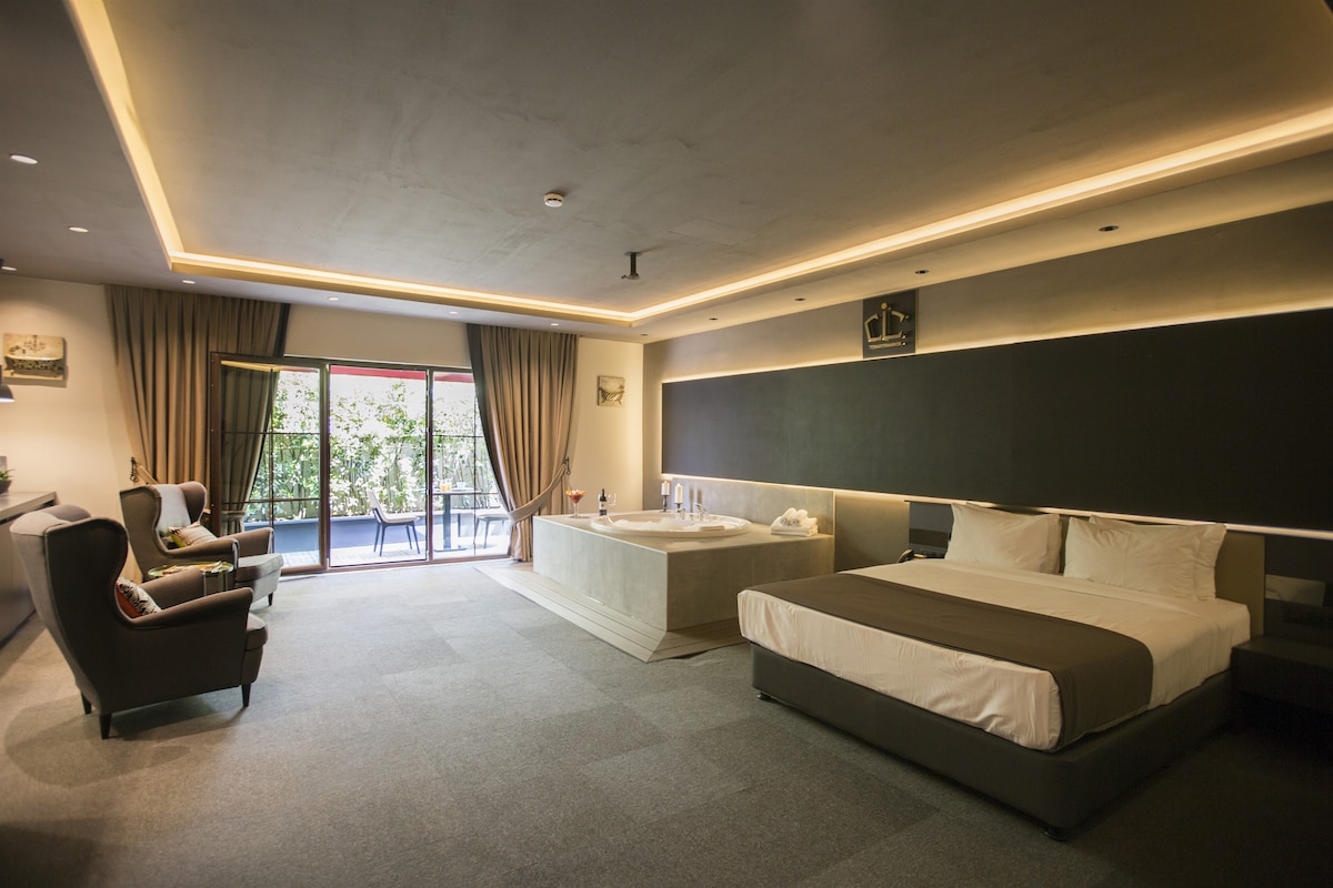 A spacious room is equipped with a large bed and a minimalist seating area featuring two armchairs. A desk is positioned nearby, while glass doors lead to a private balcony surrounded by greenery. Soft lighting enhances the modern decor and relaxed atmosphere.