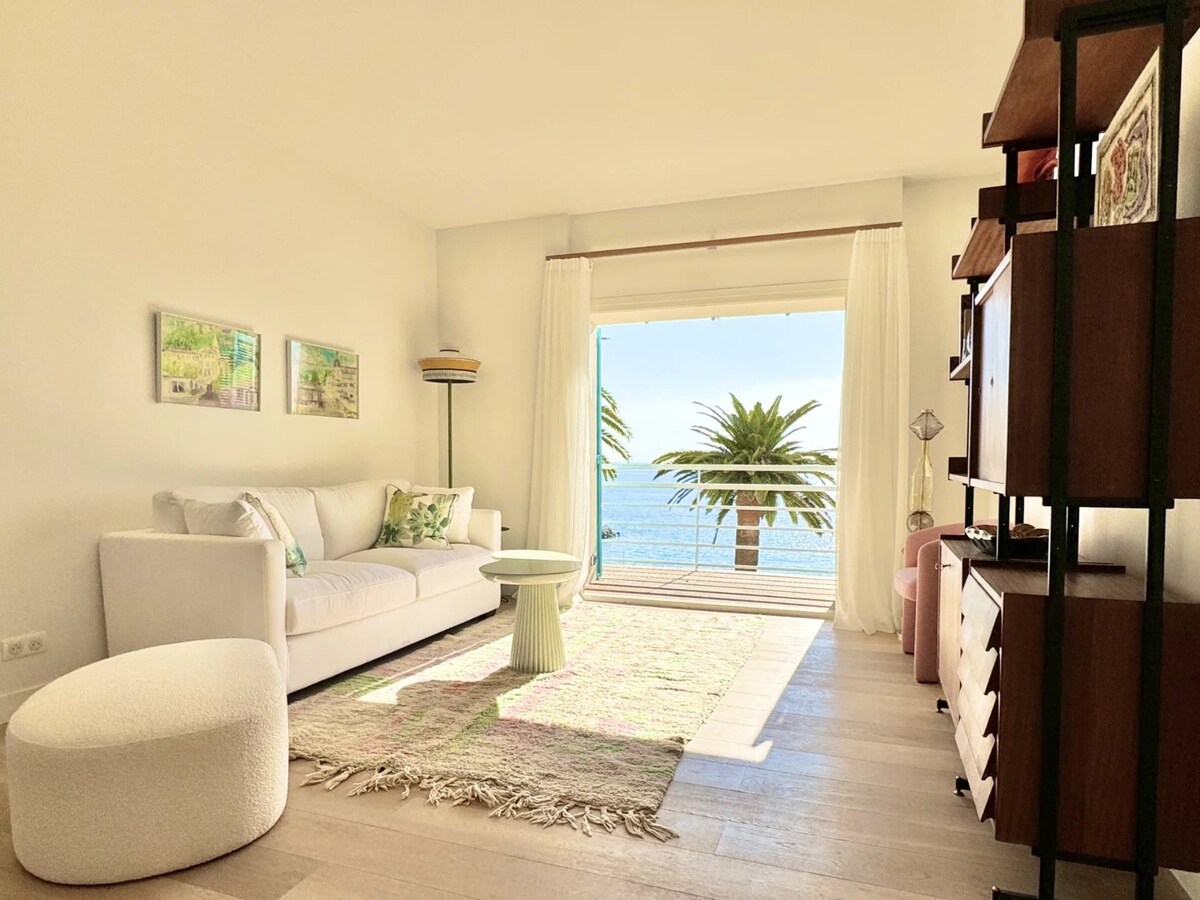 A bright living area is featured, with large sliding glass doors opening to a sea view. A comfortable white sofa and a round side table are positioned on a soft area rug. Natural light floods the space, highlighting coastal-inspired artwork on the walls.