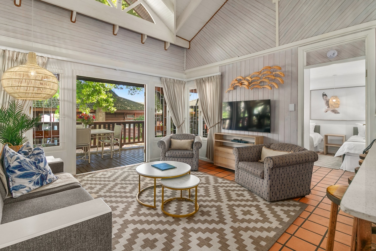 A spacious living area features large glass doors that open to a patio, bringing in natural light. Comfortable seating is arranged around a round coffee table on a patterned rug. Decor is complemented by wooden accents and greenery, creating a relaxed ambiance in the cottage.