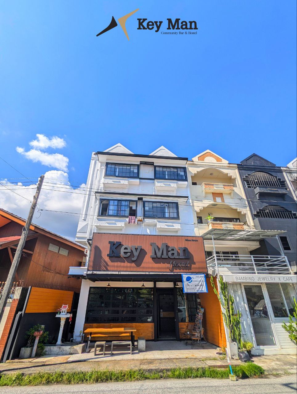 The exterior of a multi-story building is displayed, featuring a prominent sign reading 'Key Man'. Large windows are visible on each floor, with a wooden bench situated in front. The surroundings include a variety of other buildings and clear blue skies overhead.