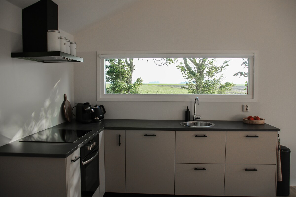 A modern kitchen features sleek cabinetry and a minimalist design. Natural light floods the space through a large window, offering a view of the surrounding fields. Essential appliances, including a stove and sink, are integrated into the layout, enhancing functionality and ease of use.