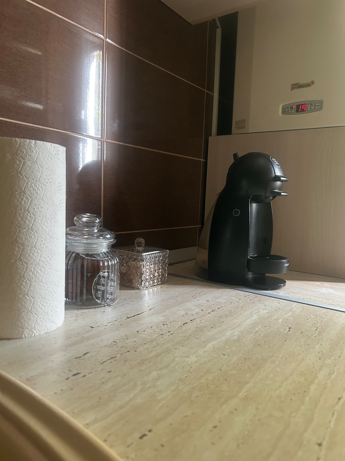 A kitchen counter is showcased, featuring a sleek coffee machine positioned beside a glass jar with a lid. A small, decorative container holds various items. The natural stone surface adds a warm tone, complemented by the dark tiles in the background.