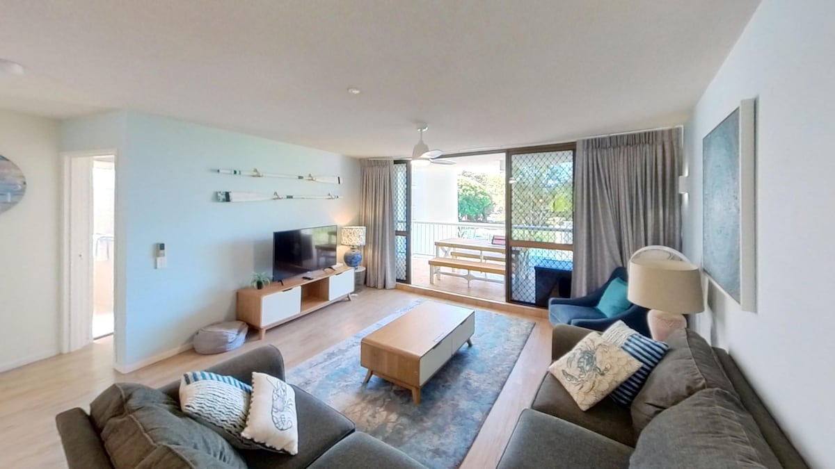 A spacious living area features comfortable seating arrangements with plush sofas and a coffee table at the center. Large glass doors provide natural light and offer views of the balcony. A television is positioned on a media console, enhancing the welcoming atmosphere.