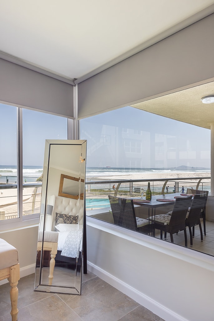 A bright corner of the living space features a large mirror reflecting the ocean view and a dining area with a table and chairs. Light streams through expansive windows, enhancing the airy atmosphere of the room.