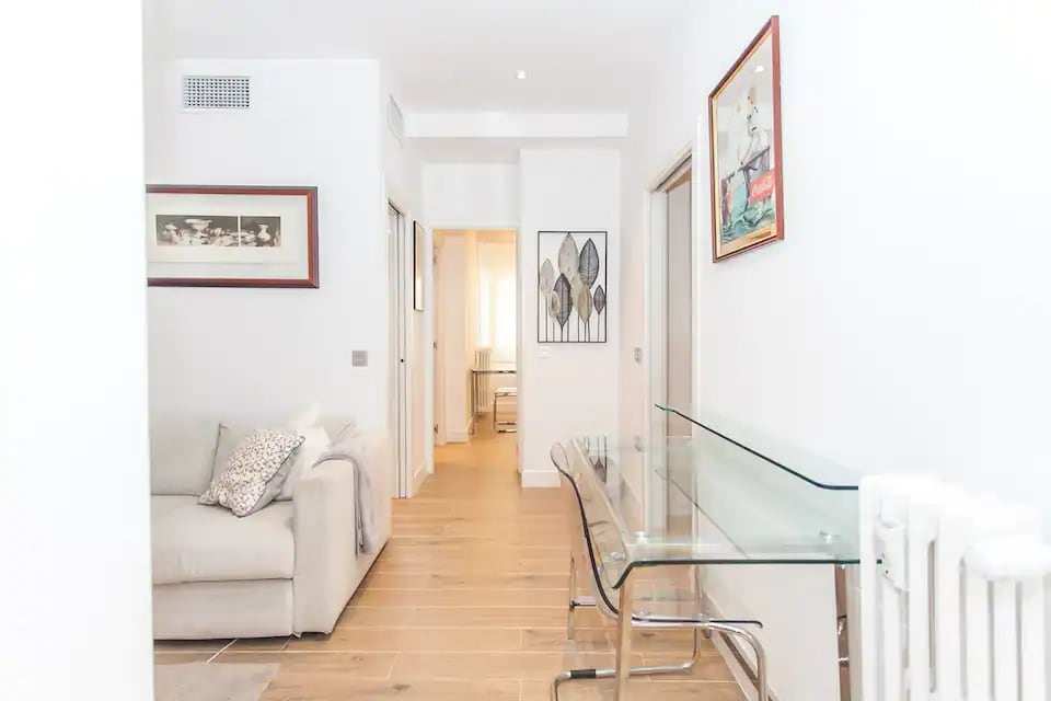 A bright and spacious common area features a light-colored sofa and a glass desk, providing a clear line of sight towards room entrances. Soft natural light filters through nearby windows, enhancing the minimalist decor that complements the overall ambiance.