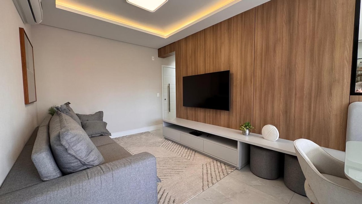 A modern living area features a comfortable gray sofa against a warm wood accent wall. A flat-screen TV is mounted on the wall, above a low-profile entertainment unit with decorative items. Natural light fills the space, creating an open and inviting atmosphere.