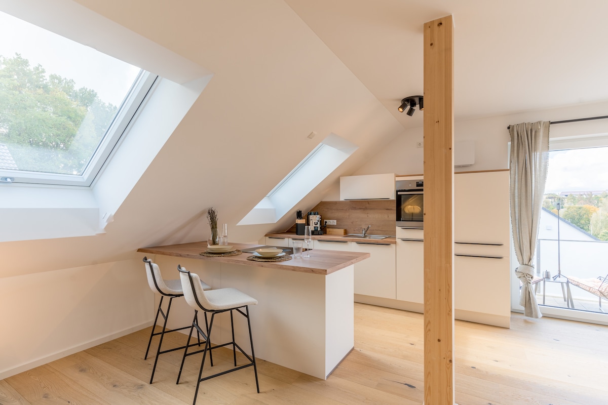 The modern kitchen is complemented by sloped ceilings and large skylights, allowing natural light to fill the space. A wooden island with seating for two is positioned near the cooking area, which features sleek cabinetry and contemporary appliances.
