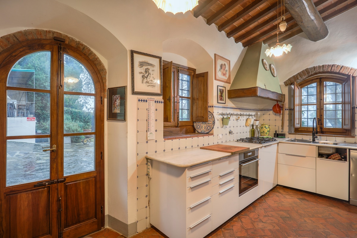 A well-equipped kitchen is featured with modern cabinetry and a spacious countertop. The room is illuminated by soft lighting, complementing the rustic charm of wooden beams and terracotta flooring. Windows provide natural light and views of the outdoors, enhancing the inviting atmosphere.