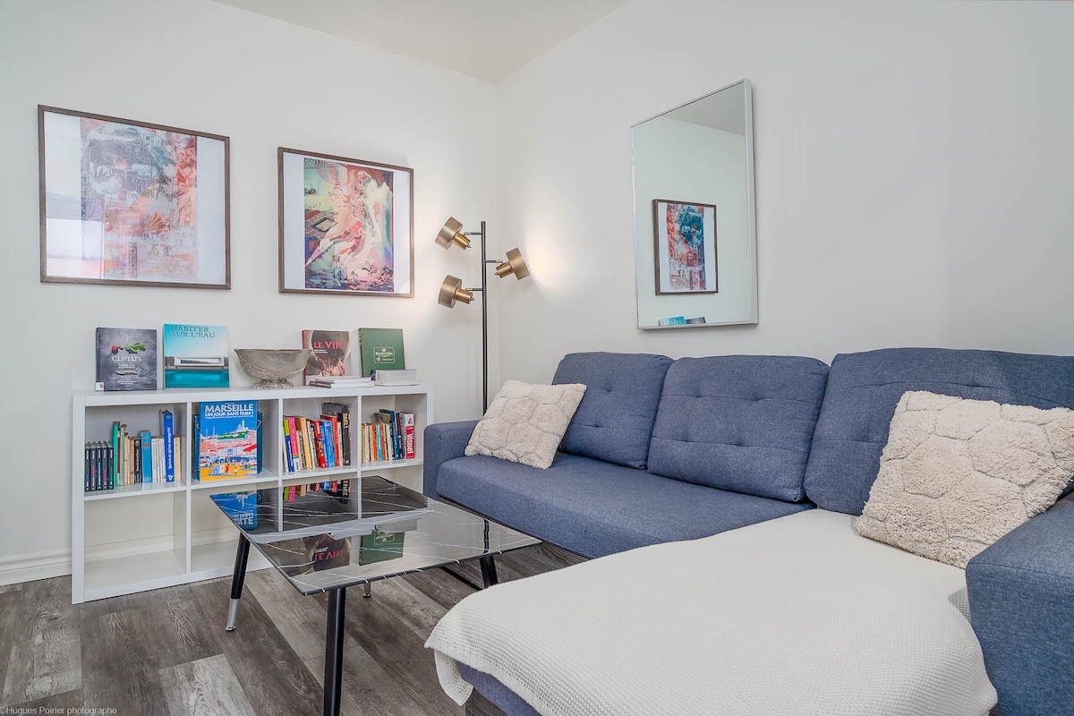 A comfortable living area features a modern gray sectional sofa adorned with soft pillows. A glass coffee table sits in front of the sofa, complemented by a white bookshelf filled with books and decorative items. Vibrant artwork is displayed on the wall, enhancing the space's character.