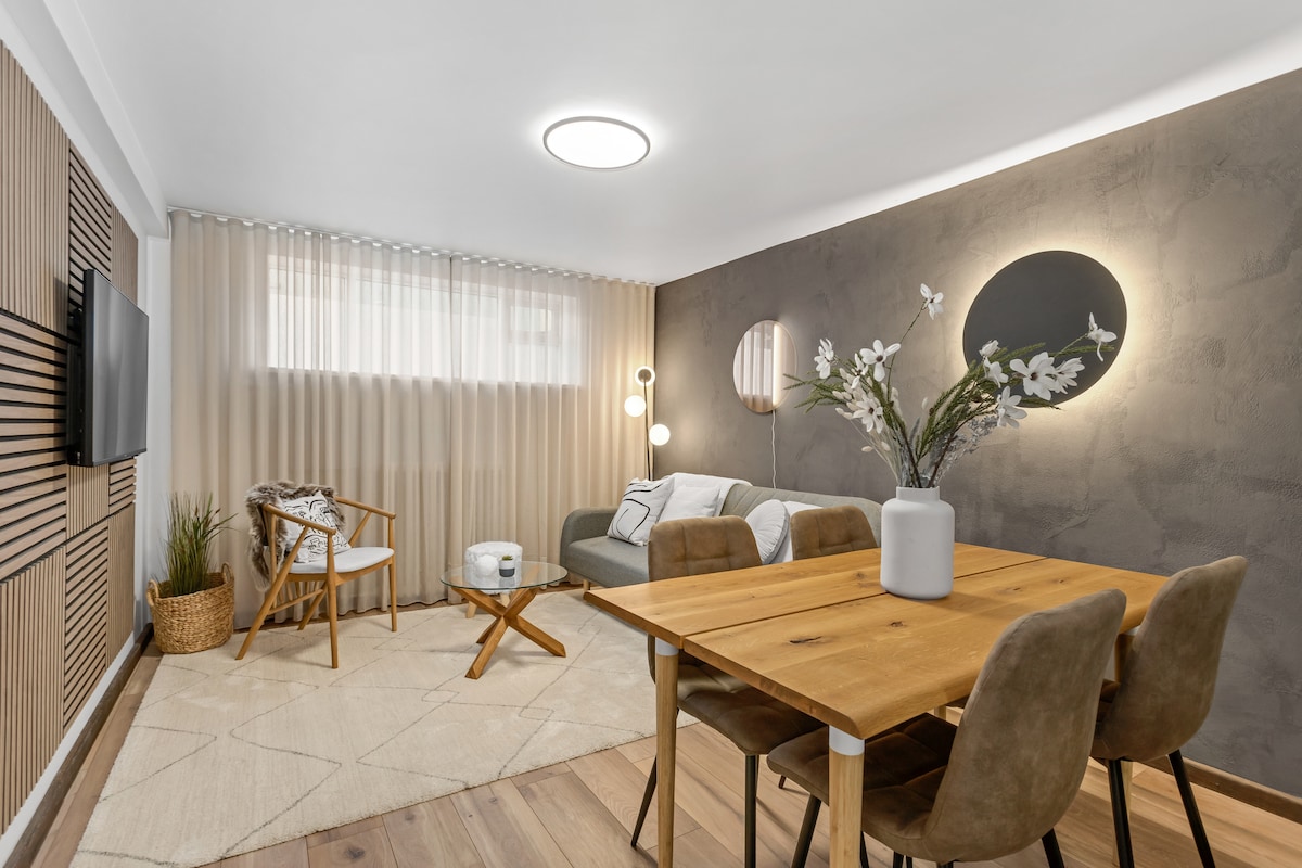 A cozy living area is furnished with a comfortable sofa and armchair, accompanied by a small table. A wooden dining table with upholstered chairs is positioned nearby. Subtle lighting is provided by wall sconces and a ceiling fixture, complementing the neutral color palette.