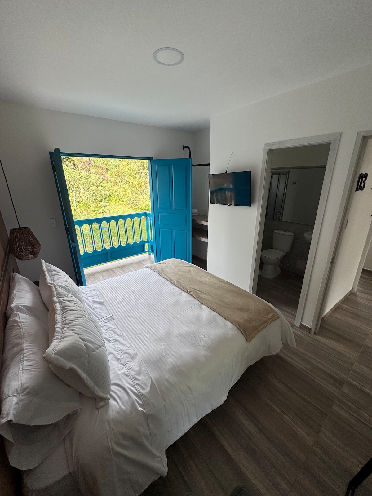 A modern bedroom features a spacious bed adorned with a light-colored blanket. Bright blue doors open to a balcony, allowing natural light to fill the room. A TV is mounted on the wall, and a doorway leads to a bathroom.