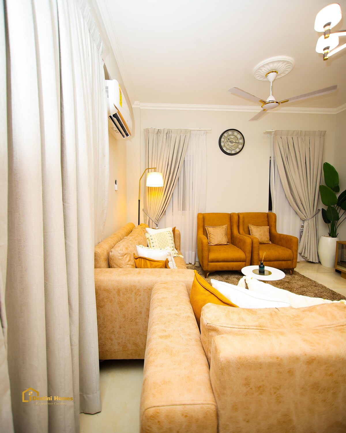 The living area is furnished with a comfortable beige sofa and two orange accent chairs. A round coffee table sits at the center. Sheer curtains frame the windows, and a ceiling fan is visible above. A green plant adds a touch of nature to the space.