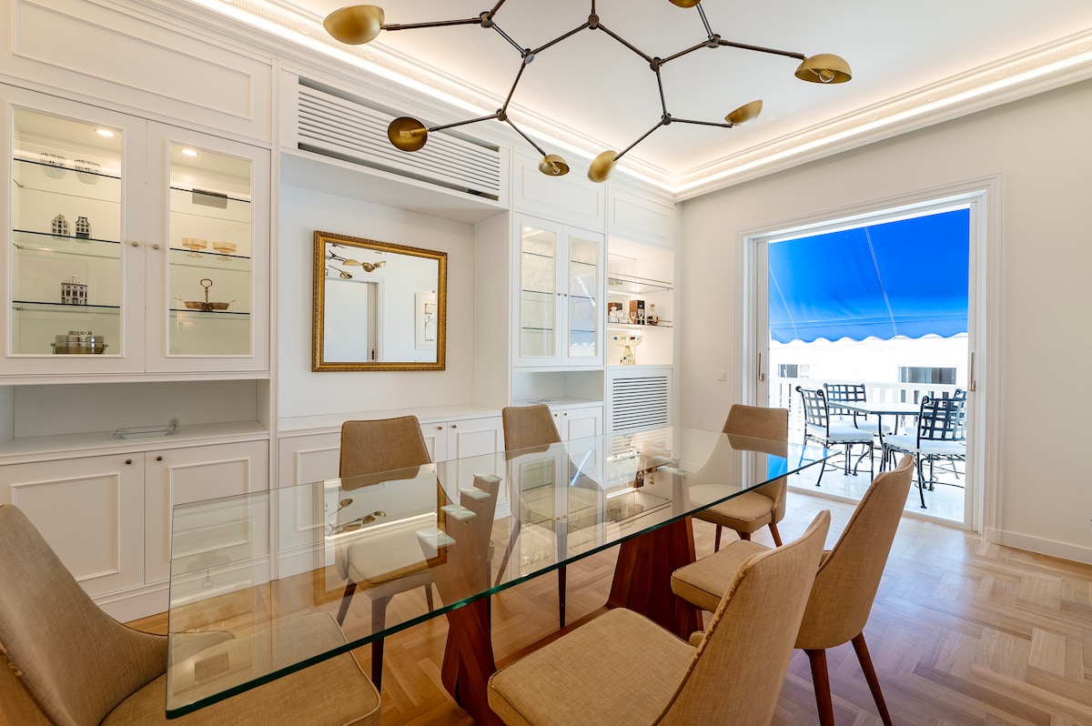 A modern dining area is depicted with a glass table surrounded by six upholstered chairs. Bright cabinetry with glass doors showcases decorative items, while a large mirror reflects the room's elegance. A sunny terrace is visible through the open door, providing outdoor seating.