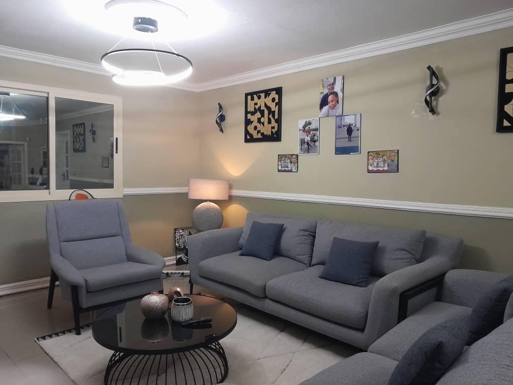 A modern living room features a circular coffee table at its center, surrounded by gray sofas with decorative cushions. A floor lamp provides soft lighting, and framed photos adorn the walls. Natural light enters through a nearby window, enhancing the inviting atmosphere.
