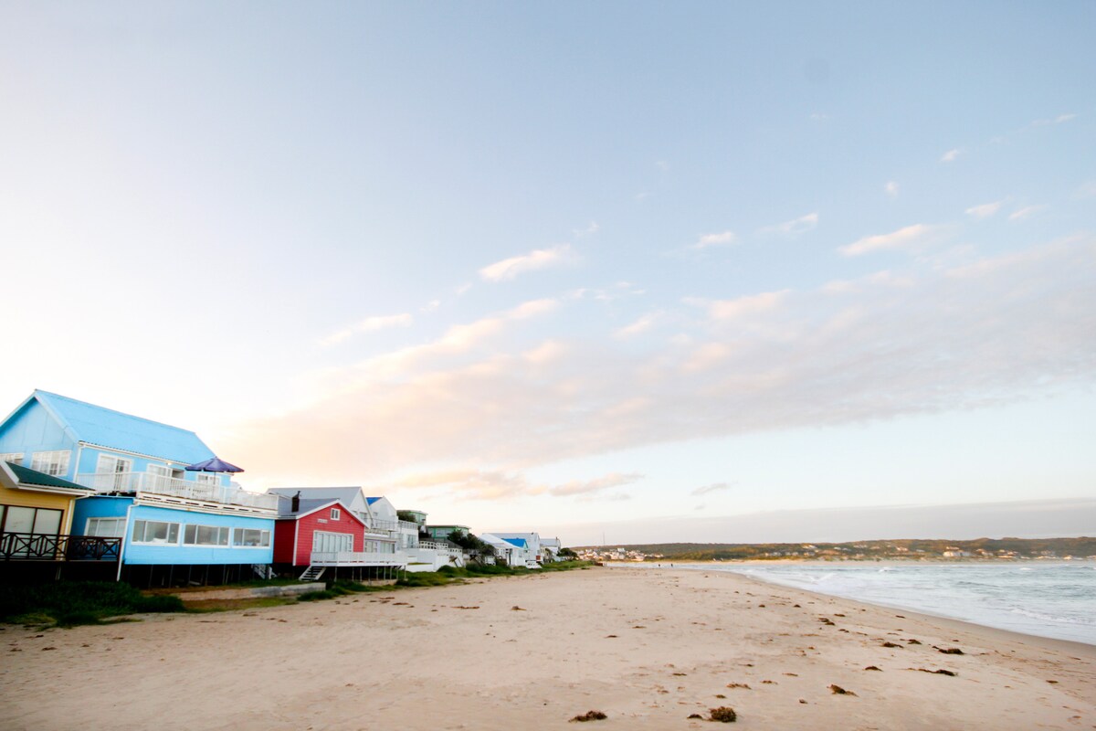 A scenic view of a sandy beach is presented, lined with colorful houses. The structures feature a variety of pastel colors, with blue and red hues standing out. The shoreline meets gentle ocean waves under a soft, cloud-streaked sky.