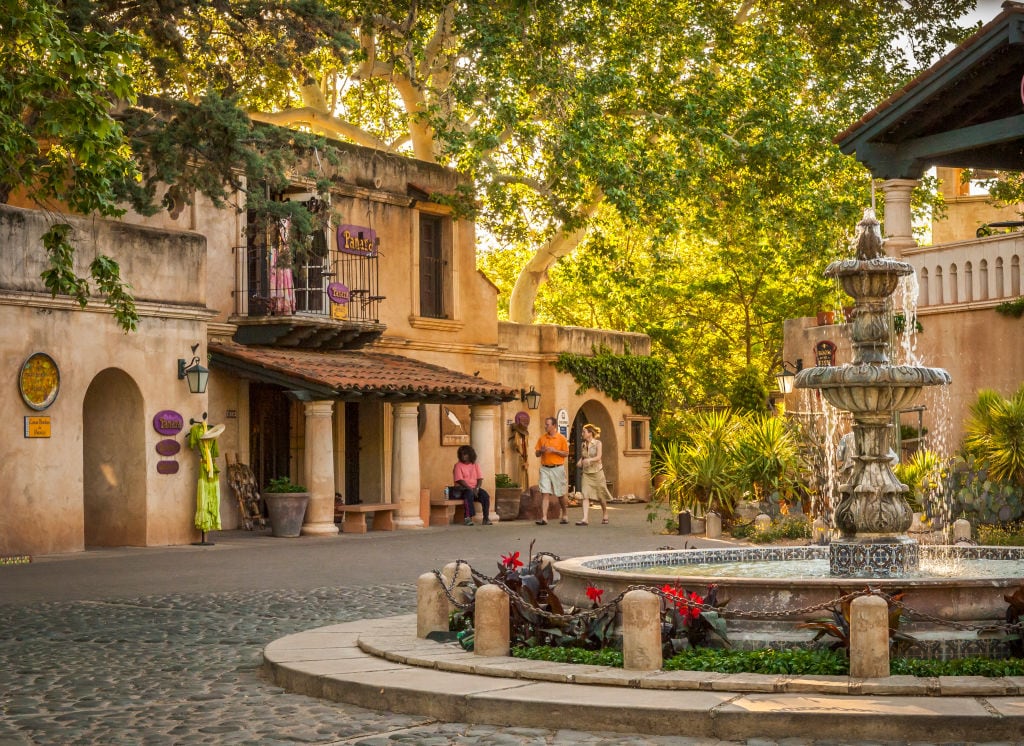 A charming courtyard is depicted, featuring a decorative fountain surrounded by lush greenery and vibrant flowers. Historic-style buildings with balconies are visible, complemented by cobblestone pathways. Soft sunlight filters through the trees, creating a serene outdoor atmosphere.