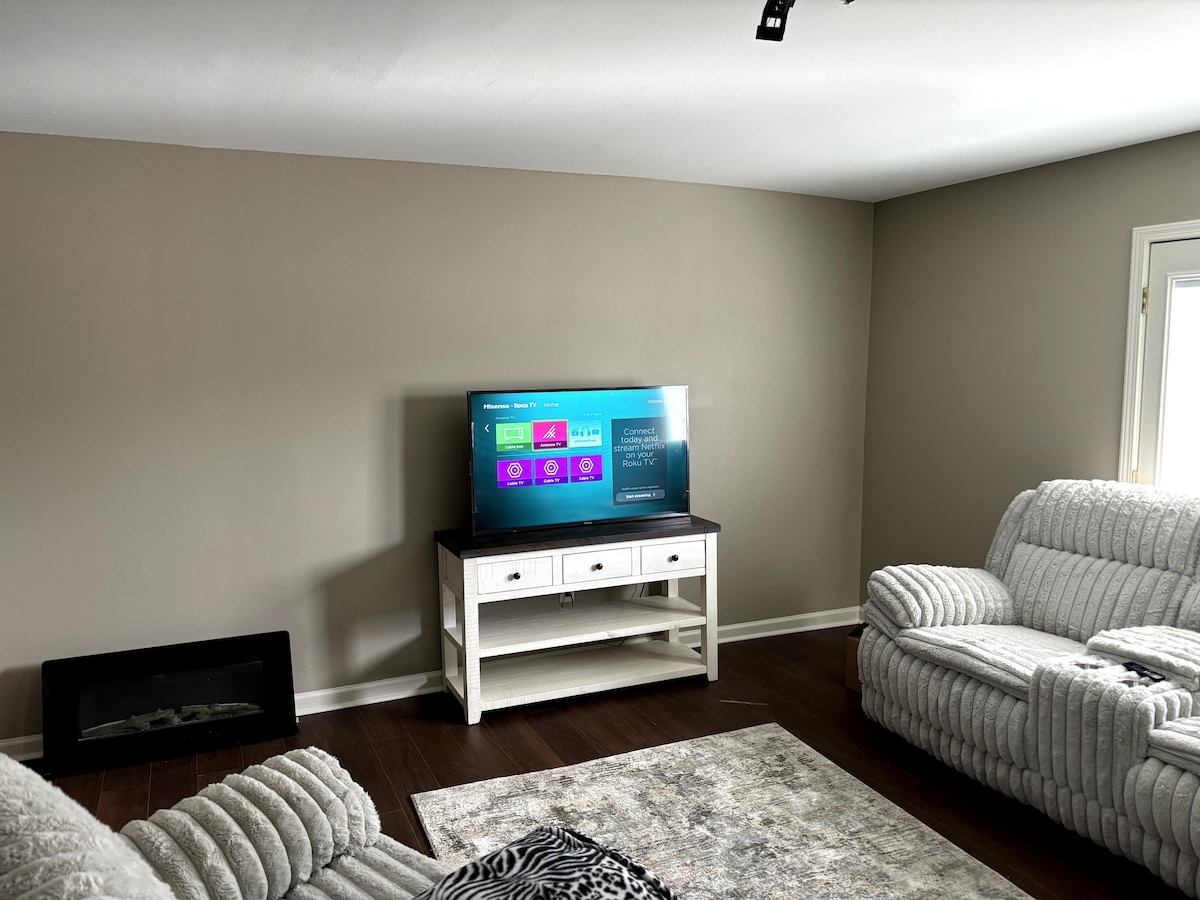 A comfortable living room is presented with two plush sofas positioned around a stylish area rug. A television mounted on the wall provides entertainment options, while a classic console table offers additional decor. Natural light enters through the nearby windows, enhancing the space.