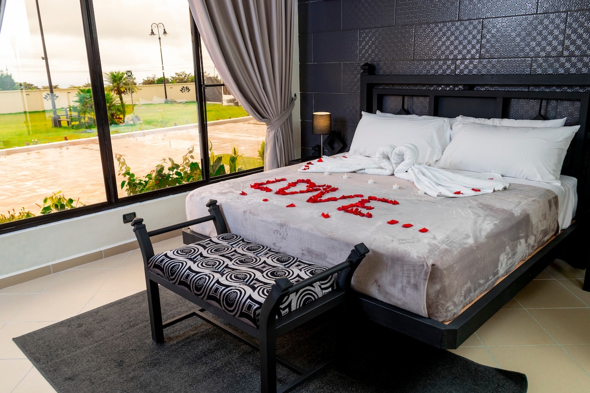 A spacious bed is adorned with rose petals forming the word 'LOVE.' A stylish bench with a geometric pattern sits at the foot of the bed. Large windows offer a view of the outdoors, complemented by soothing light filtering through the elegant curtains.