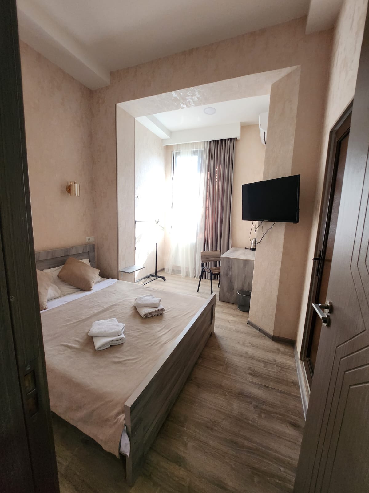 A well-arranged sleeping area features a spacious bed with neutral linens and decorative pillows. A small TV is mounted on the wall, while a chair and table provide a functional workspace. Natural light enters through the window, which is dressed in soft drapes.