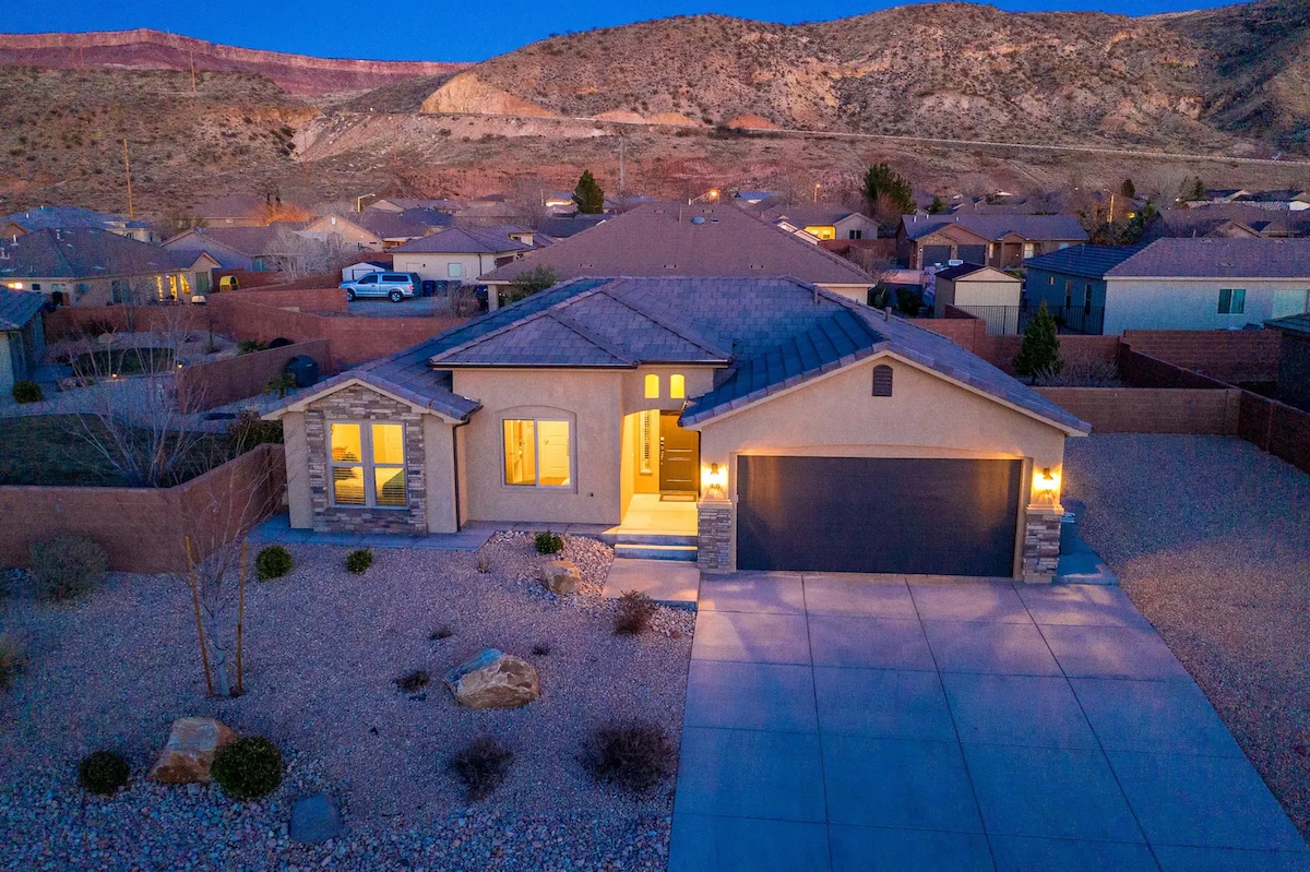 An aerial view of a modern single-story home is captured at dusk, showcasing warm exterior lighting. A concrete driveway leads to an attached garage, while neatly landscaped gravel surrounds the home. Scenic hills appear in the background, illustrating the tranquil setting.