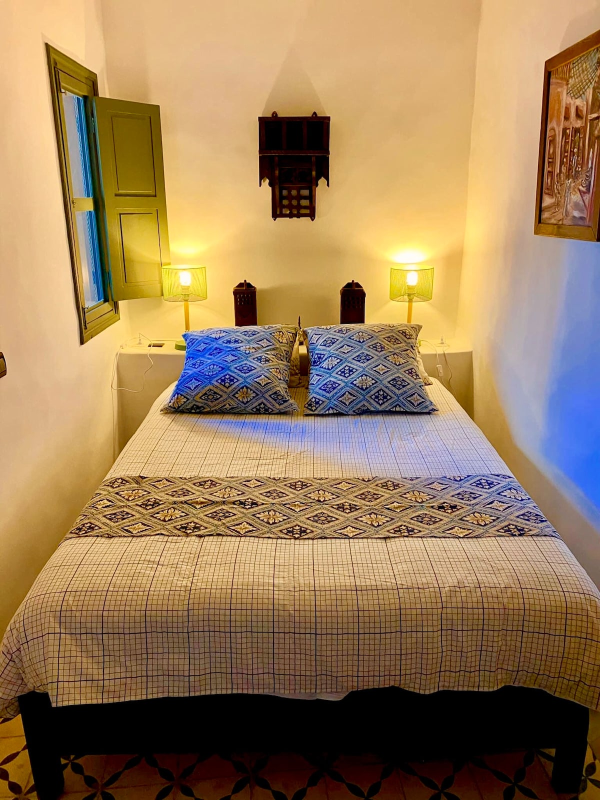 A cozy bedroom is displayed, showcasing a spacious bed adorned with patterned pillows and a matching blanket. Soft lighting is provided by two bedside lamps, creating a warm glow. A window with green shutters allows for natural light, and artwork is positioned on the wall.