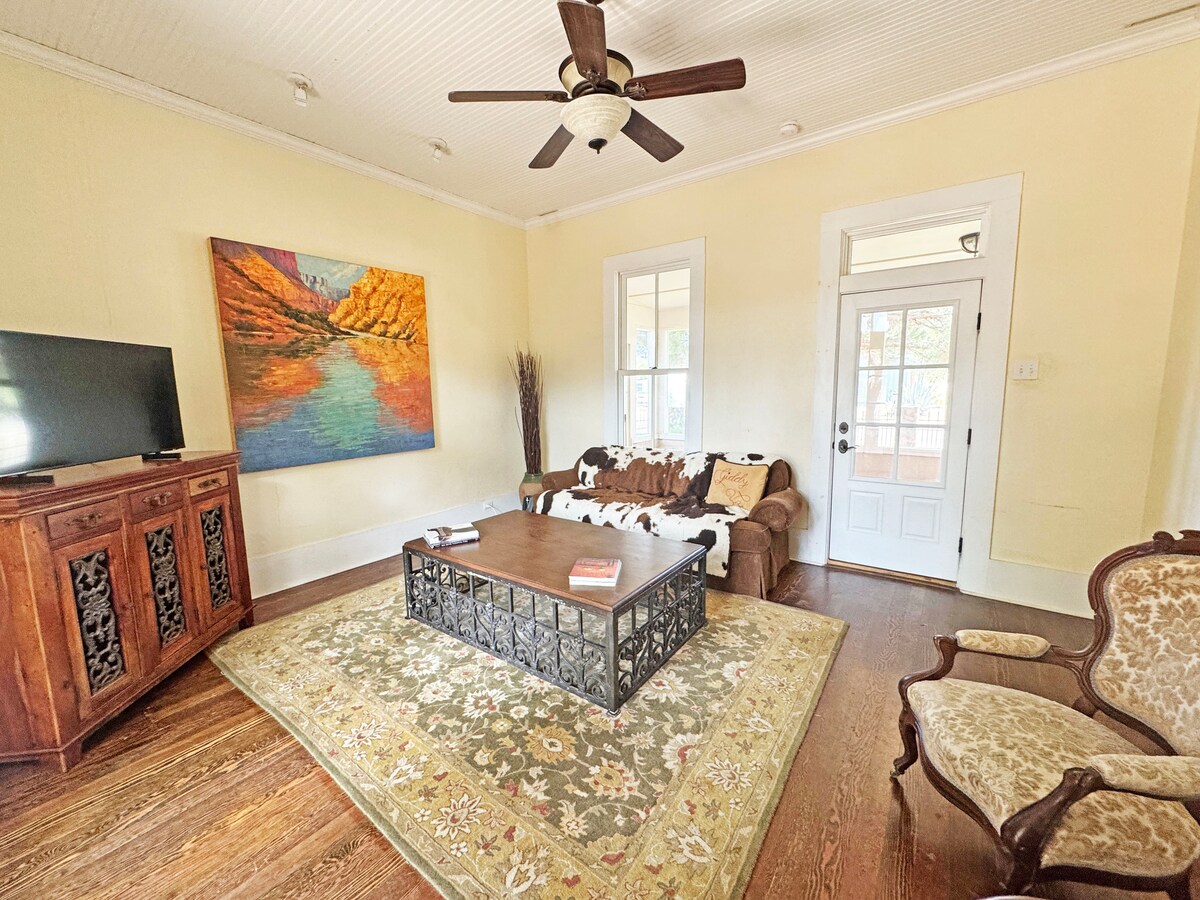 The living area features a wooden coffee table centered on a patterned rug, complemented by a vintage side chair and a TV unit. A large artwork adorns the wall, while natural light enters through the door and windows, illuminating the warm yellow walls.