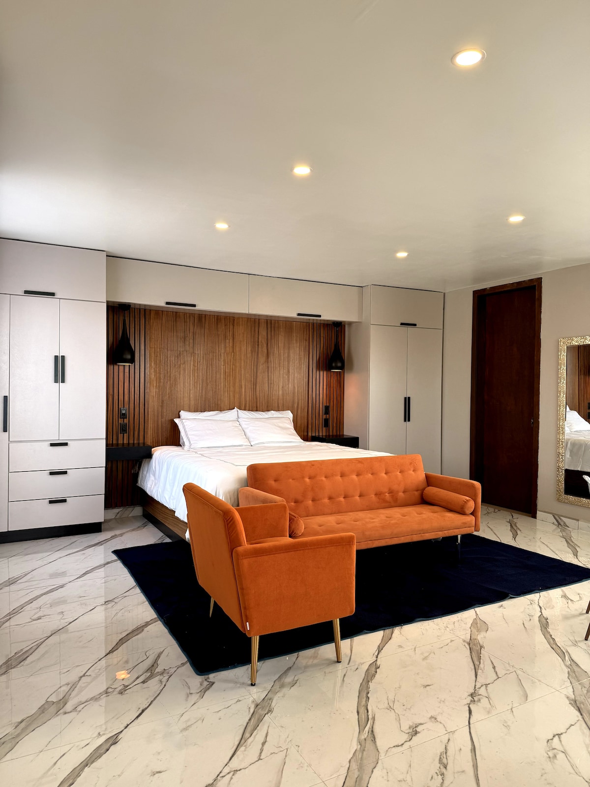 A spacious bedroom features a large, comfortable bed, complemented by warm wood accents on the wall. An orange loveseat and matching armchair are positioned on a dark rug, adding a pop of color to the room. The marble flooring reflects the soft, overhead lighting.
