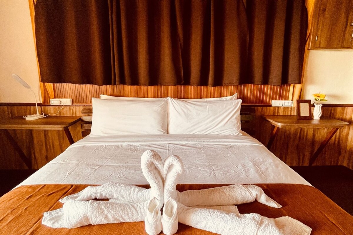 A spacious bed is adorned with crisp white linens, featuring elegantly folded towel swans at the foot. Dark curtains frame the window, allowing soft light to partially illuminate the room, while wooden side tables are positioned on either side.