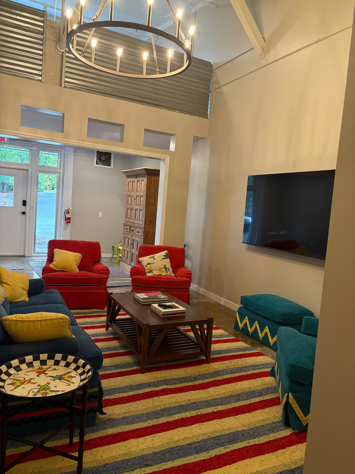 A spacious living area is furnished with two vibrant red chairs and a light blue couch. A large flat-screen TV is mounted on the wall, opposite a striped area rug. A wooden coffee table holds decorative items, while natural light enters through large doors.