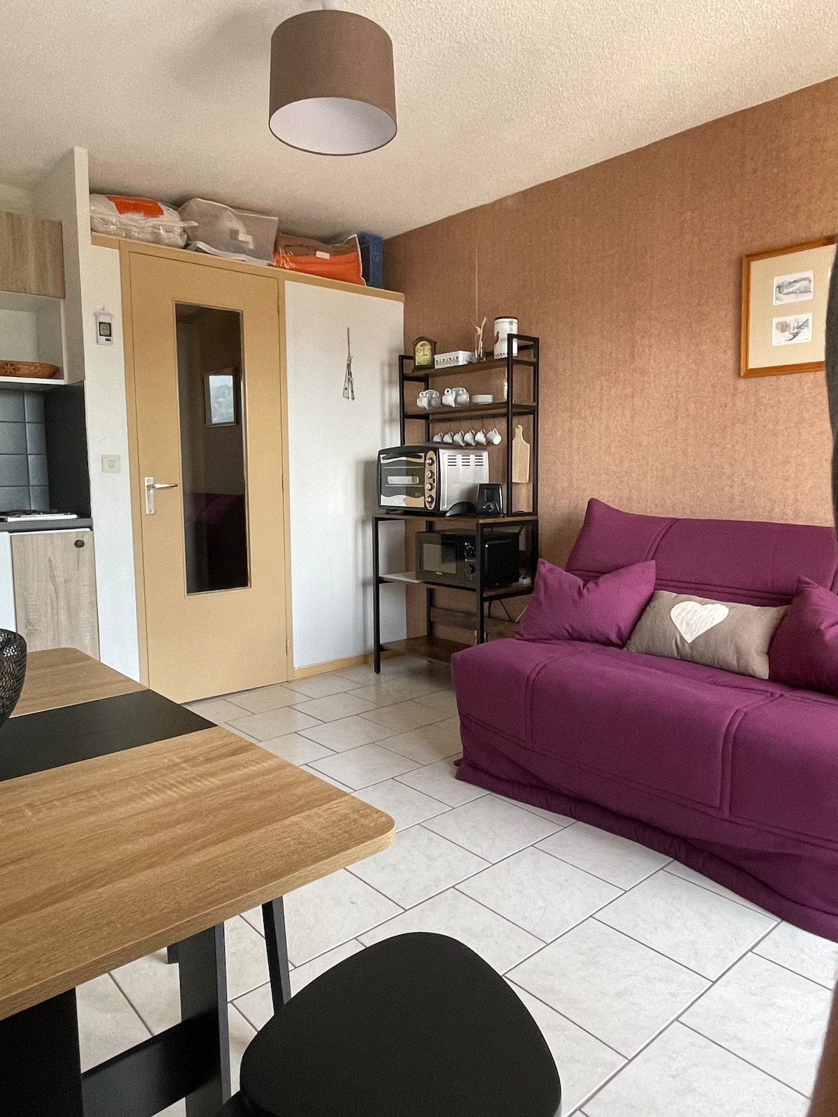 The living area includes a purple sofa with cushions, positioned near a sleek wooden dining table and black chairs. A tall bookshelf holds kitchen essentials, with a microwave nearby. Neutral-toned walls are complemented by natural light filtering through a window, enhancing the cozy atmosphere.