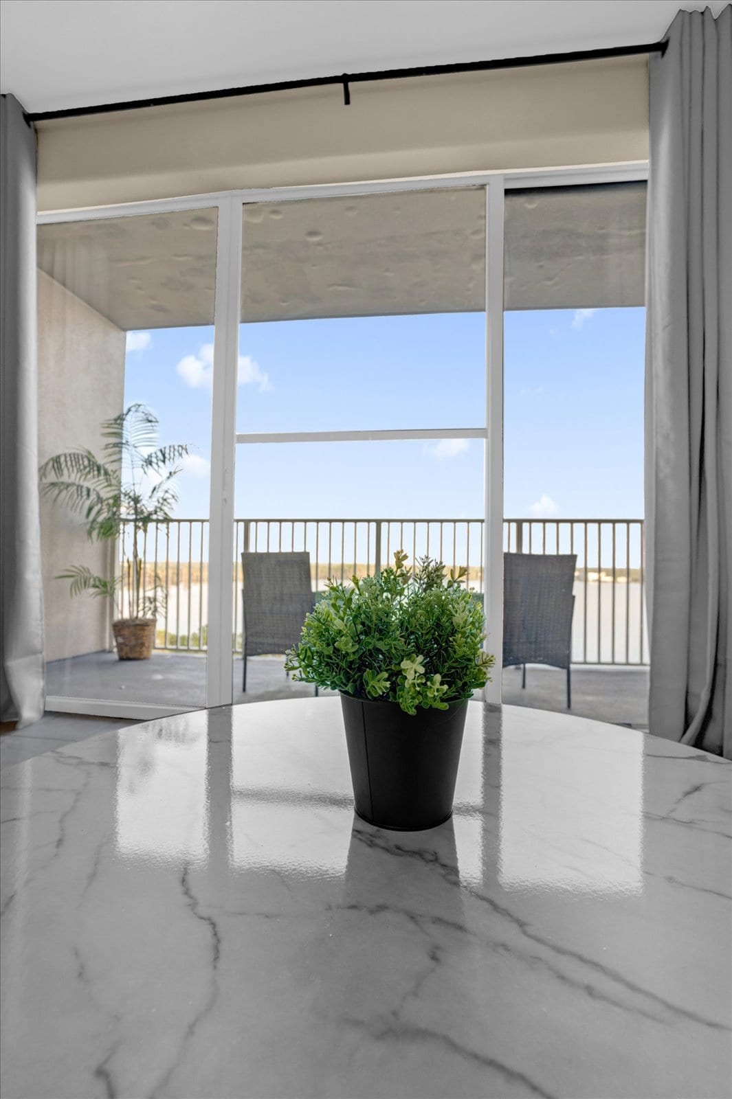 A modern coffee table featuring a small potted plant sits in the foreground, with large sliding glass doors in the background leading to a balcony. The serene outdoor view showcases a clear sky, while two chairs positioned on the balcony are visible.