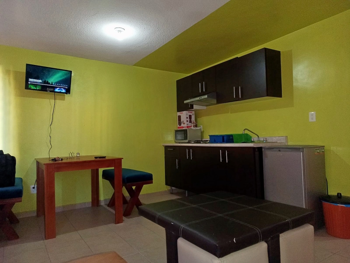 A compact living area features a small wooden table with two upholstered chairs and a dark countertop. A microwave and a refrigerator are present in the kitchen area, with colorfully organized dishes and utensils. Light yellow walls enhance the bright and functional space.