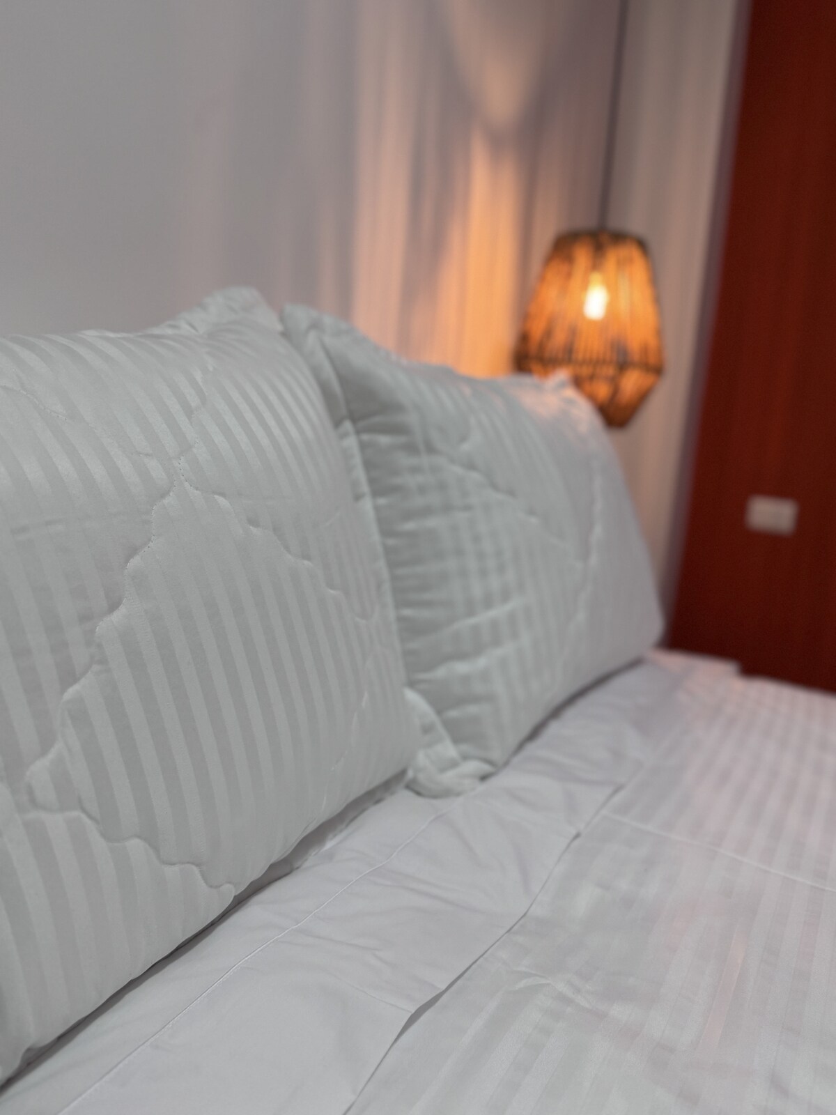 Plush white pillows are arranged neatly against a headboard, complemented by a warm light source from a woven lamp in the background. The bedding features a clean, textured design, contributing to a relaxing ambiance.